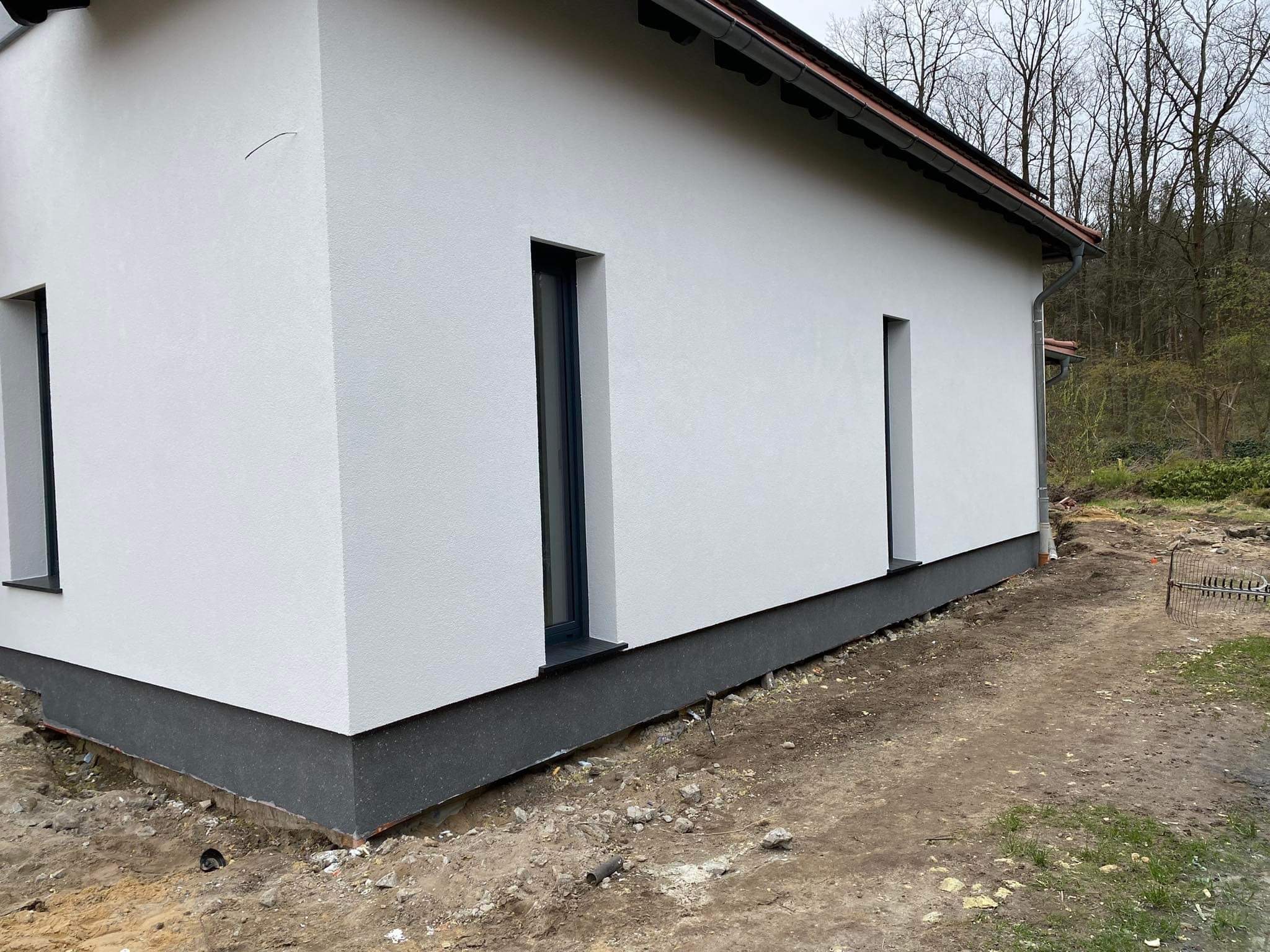 Nowoczesny dom jednorodzinny z białą elewacją o fakturze baranka i ciemnoszarą podmurówką, widoczne okna z ciemnymi ramami i fragment dachu z rynnami, teren wokół domu w trakcie prac wykończeniowych.
