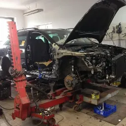 Uszkodzony samochód Audi na ramie naprawczej w warsztacie, z otwartą maską i zdemontowanymi elementami karoserii, widoczne narzędzia na ścianie.