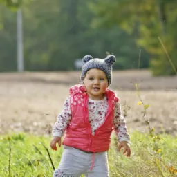 Uśmiechnięta, mała dziewczynka w różowej kamizelce i czapce z uszkami stoi na trawie w słoneczny dzień.