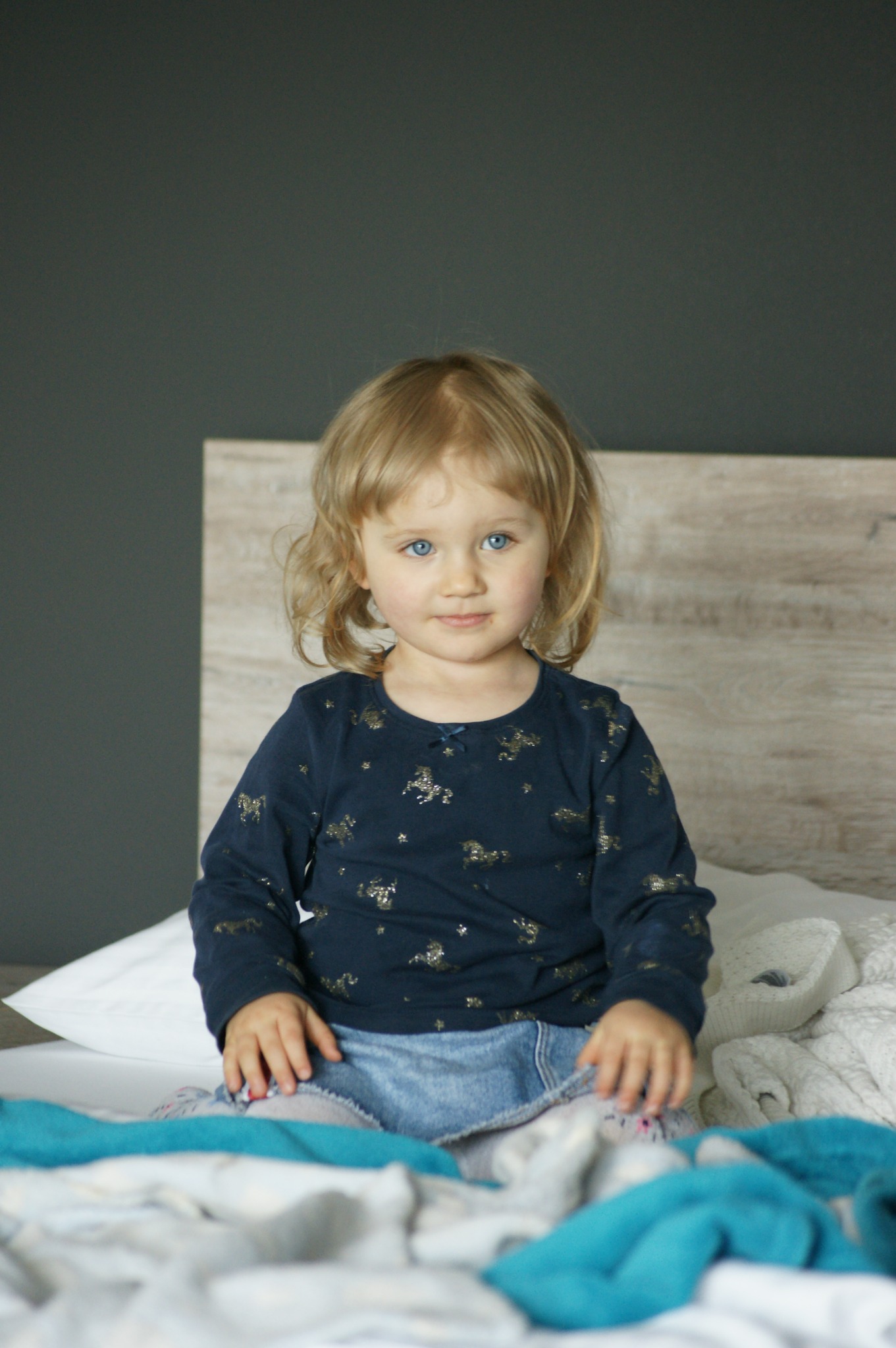 Portret uroczej dziewczynki z blond włosami i niebieskimi oczami, siedzącej na łóżku z turkusowym kocem, ubrana w granatową bluzkę w złote koniki i dżinsową spódniczkę