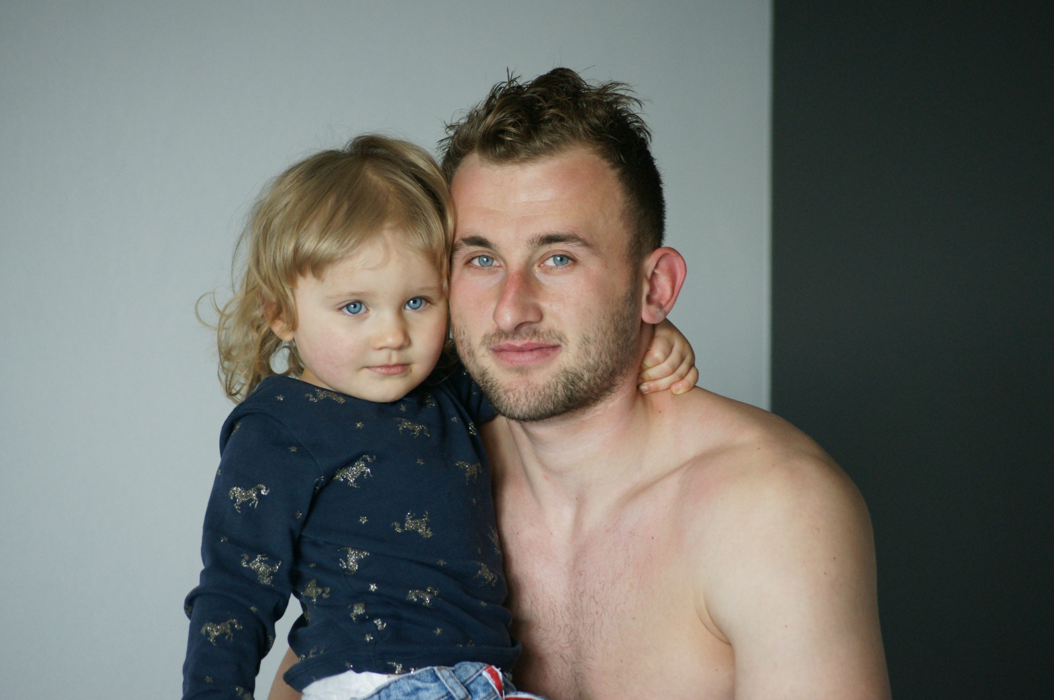 Portret ojca z córeczką o niebieskich oczach, dziewczynka w bluzce w złote koniki, mężczyzna bez koszuli, neutralne tło.