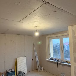 Mariusz Szafarski - Pokój w trakcie montażu płyt kartonowo-gipsowych na ścianach i suficie, widoczne narzędzia i materiały budowlane, surowy stan wykończenia, okno z widokiem na zewnątrz.
