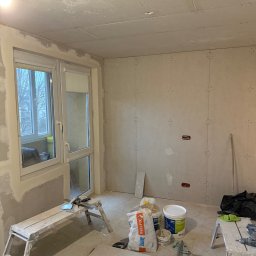 Mariusz Szafarski - Pokój w trakcie prac wykończeniowych: ściany obłożone płytami gipsowo-kartonowymi, widoczne puszki elektryczne, worki z gładzią, drabina malarska i narzędzia.