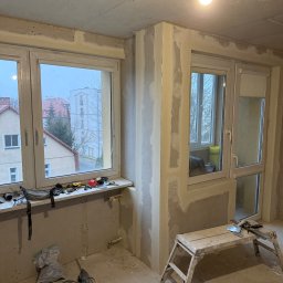 Mariusz Szafarski - Wnętrze w trakcie remontu: ściany pokryte płytami kartonowo-gipsowymi, częściowo wyszpachlowane, narzędzia leżące na parapecie okna, drabina malarska w rogu pomieszczenia.