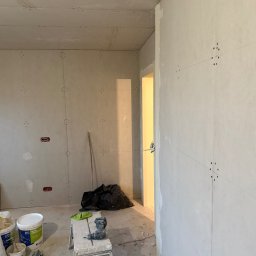 Mariusz Szafarski - Wnętrze w trakcie montażu płyt kartonowo-gipsowych na ścianach i suficie, widoczne łączenia płyt, szpachla, drabina, wiertarka i worki z odpadami w tle.