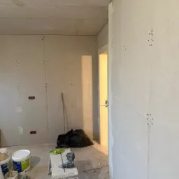 Wnętrze w trakcie montażu płyt kartonowo-gipsowych na ścianach i suficie, widoczne łączenia płyt, szpachla, drabina, wiertarka i worki z odpadami w tle.