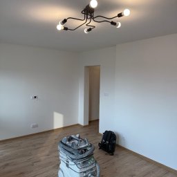 Mariusz Szafarski - Wnętrze pokoju po remoncie z nową podłogą, świeżo pomalowanymi ścianami i nowoczesną lampą sufitową; widoczny odkurzacz przemysłowy i plecak.