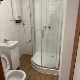 Mariusz Szafarski - Nowoczesna, mała łazienka z kabiną prysznicową z zaokrąglonymi szybami, białą umywalką z szafką, wiszącym bojlerem i toaletą z otwartą klapą, wykończona białymi płytkami i brązowymi panelami...