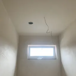 Ściany i sufit świeżo po nałożeniu gładzi szpachlowej, widoczne przewody elektryczne wystające z sufitu i otwór na oświetlenie, małe okno z widokiem na jasne światło dzienne.