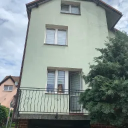 Dwupiętrowy dom z zieloną elewacją, balkonem z kutą balustradą i otwartym oknem z żaluzjami, widziany z dołu pod kątem.