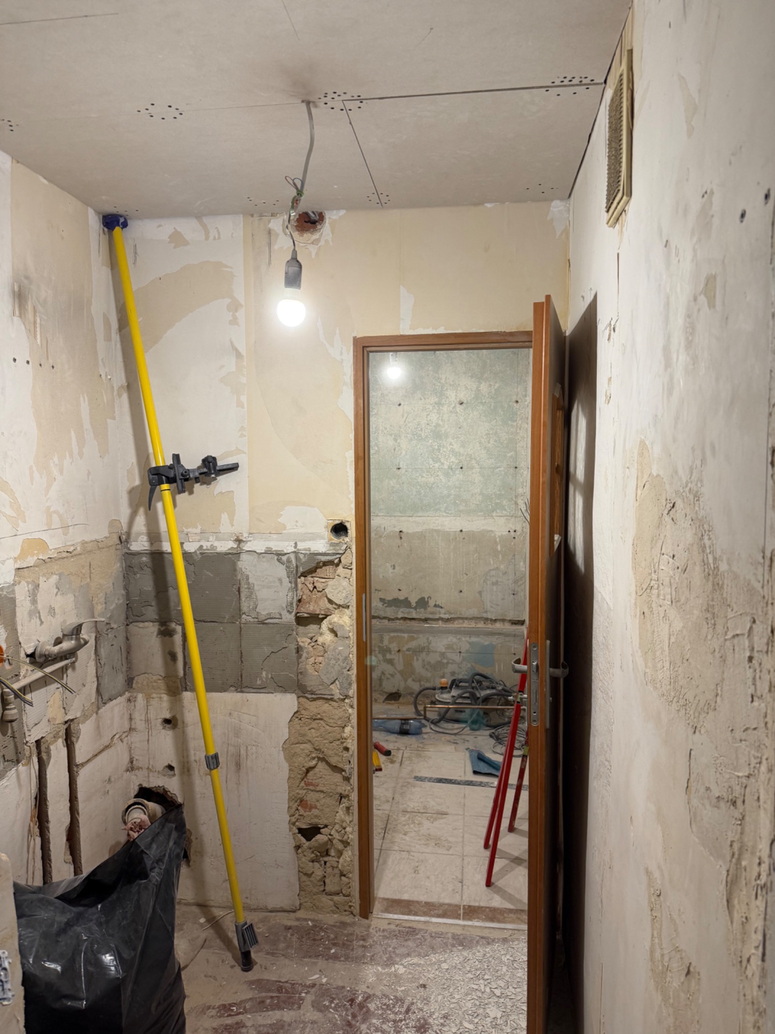 Wnętrze łazienki w trakcie demontażu, widoczne odsłonięte ściany, sufit z płyt kartonowo-gipsowych, żółty rozpierak teleskopowy i narzędzia w pomieszczeniu obok.