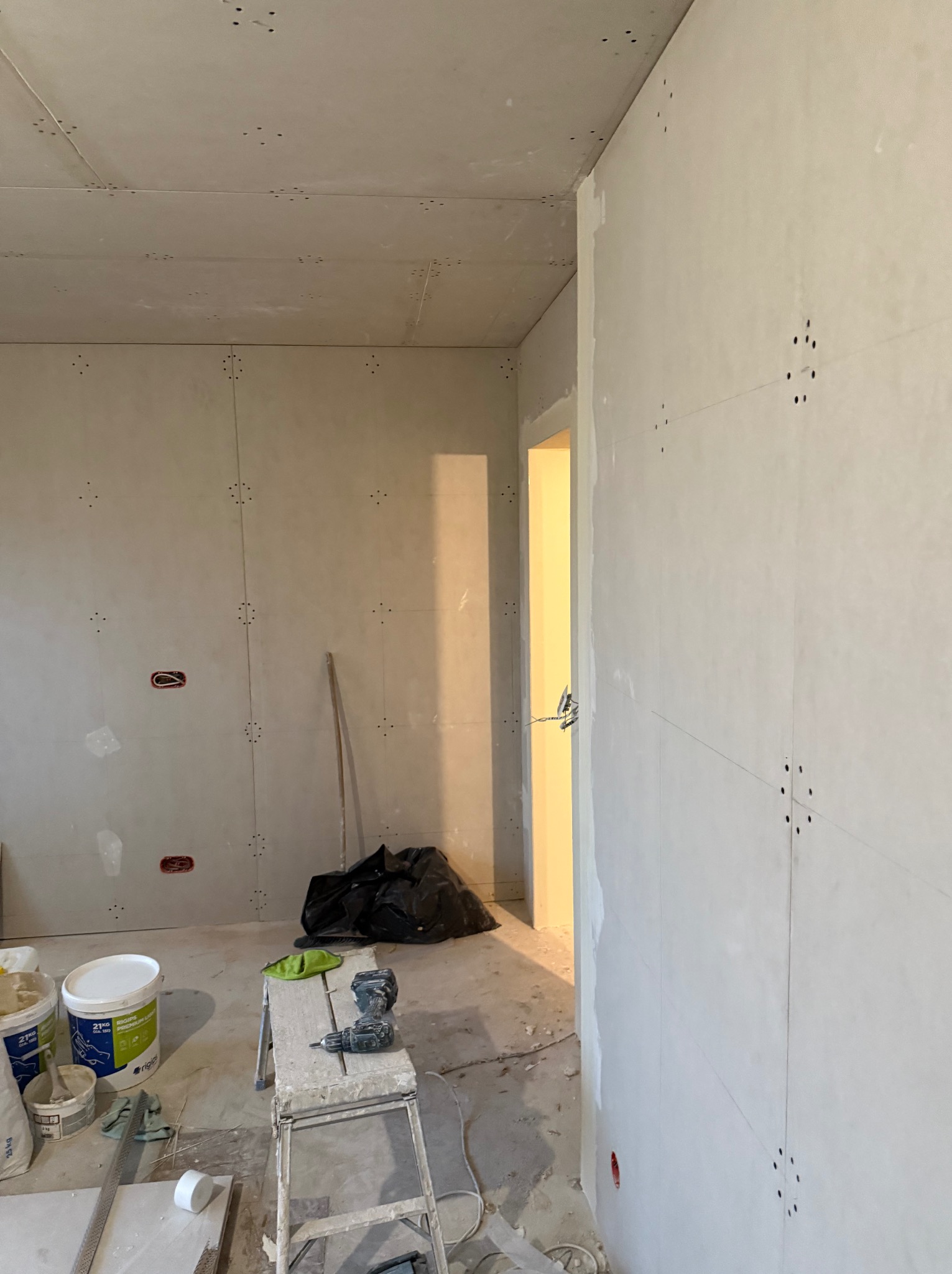 Wnętrze w trakcie montażu płyt kartonowo-gipsowych na ścianach i suficie, widoczne łączenia płyt, szpachla, drabina, wiertarka i worki z odpadami w tle.