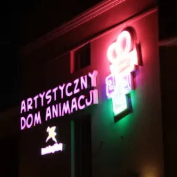 Neonowy szyld na budynku z napisem 'Artystyczny Dom Animacji' oraz symbolem kamery filmowej i logotypem firmy, oświetlone w nocy.