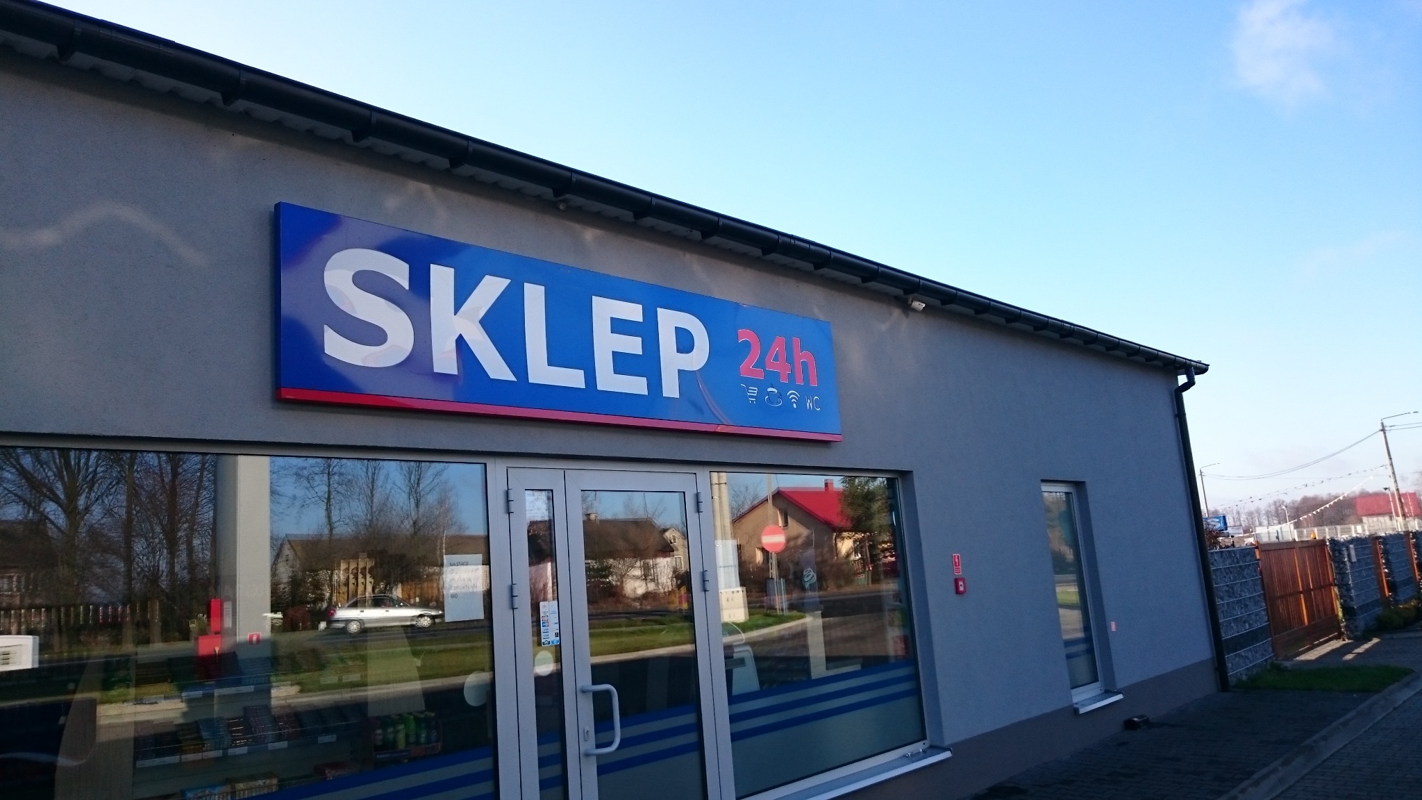 Elewacja budynku sklepu z dużym szyldem 'SKLEP 24h' w kolorach białym i czerwonym na niebieskim tle, widoczne okna i drzwi wejściowe z refleksami otoczenia.