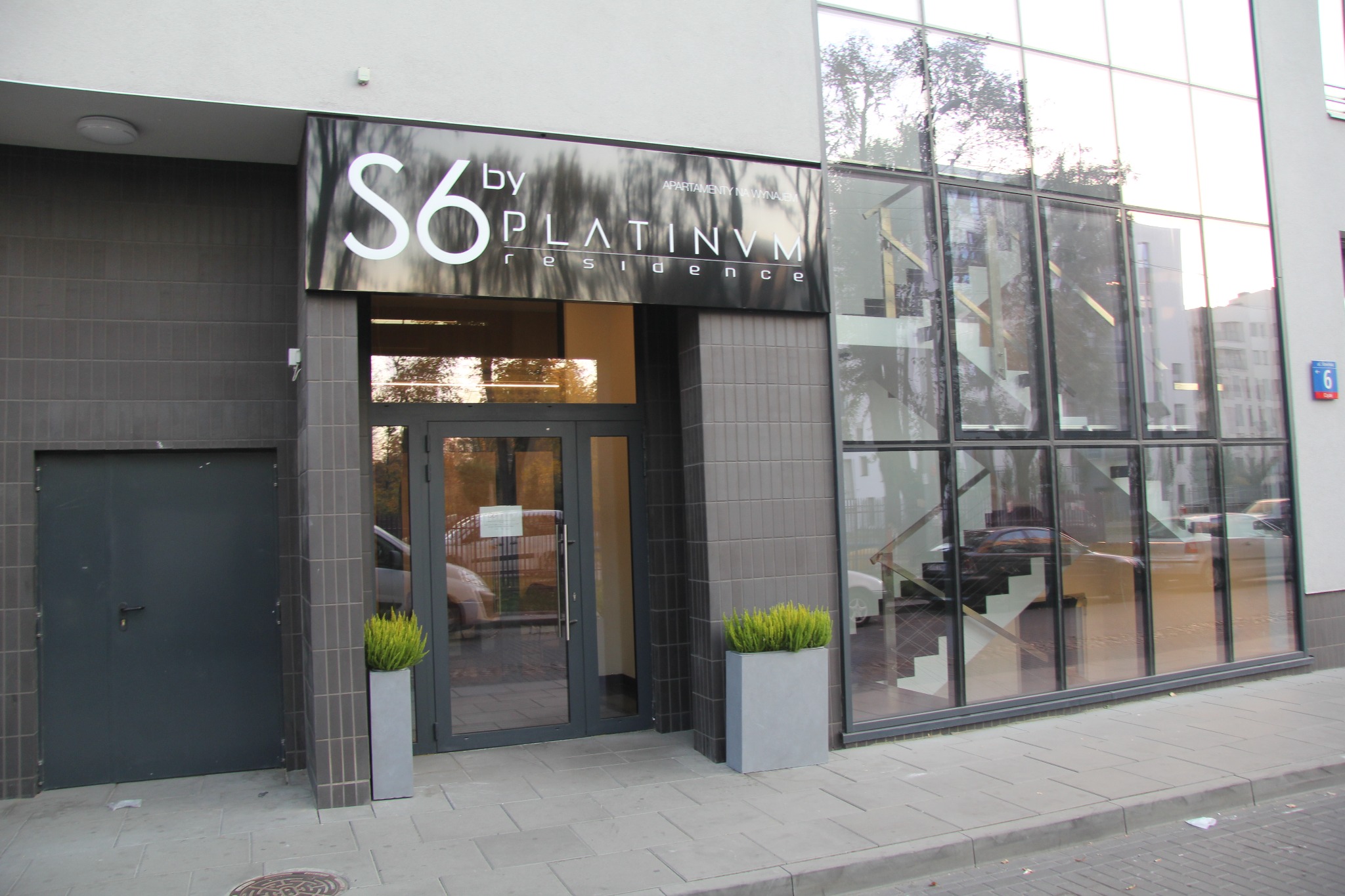 Wejście do budynku z szyldem 'S6 by PLATINVM residence' nad drzwiami i dużymi oknami z widocznymi schodami wewnątrz.