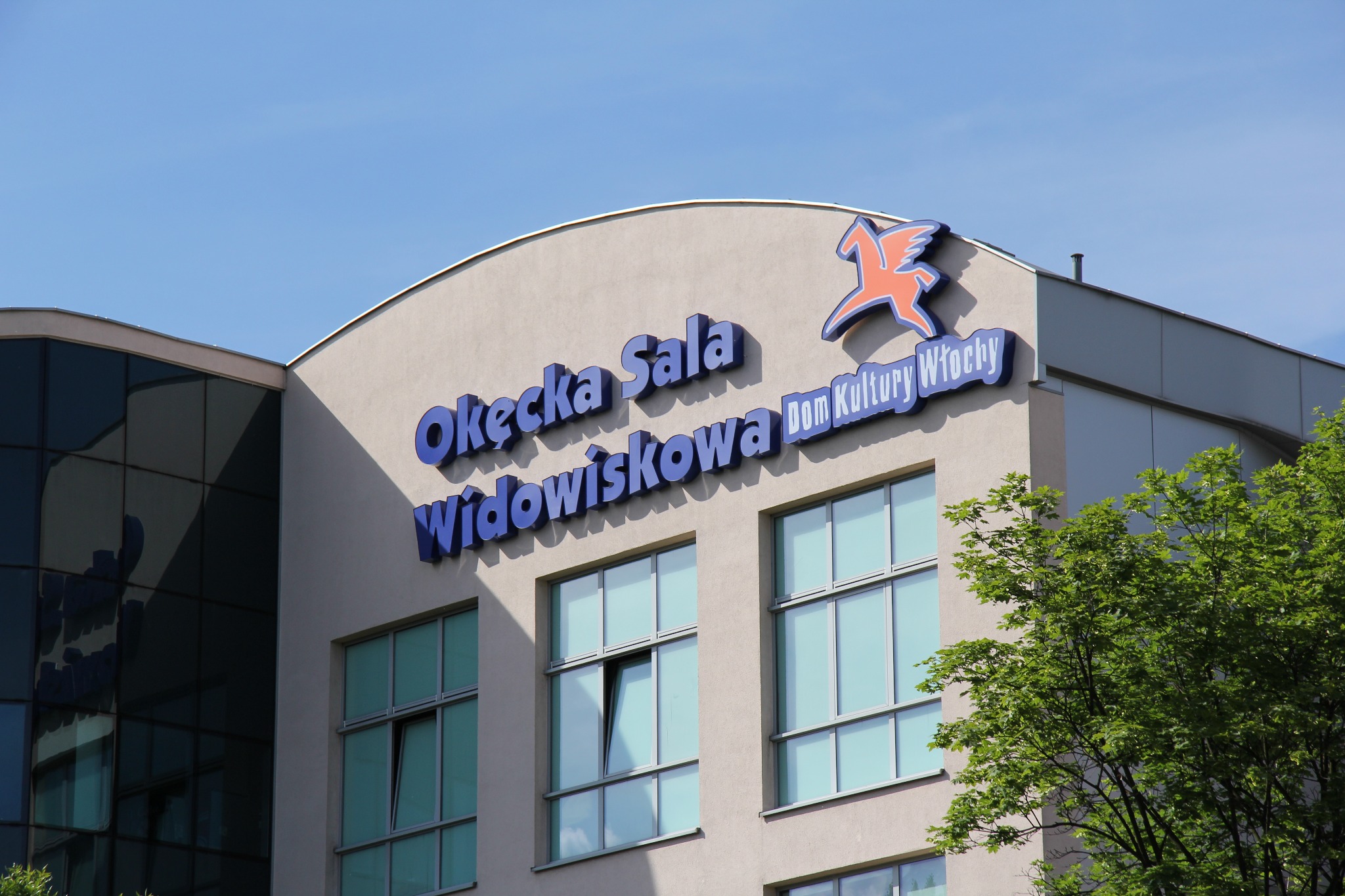 Elewacja budynku Okęcka Sala Widowiskowa Dom Kultury Włochy z niebieskim napisem i pomarańczowym logo pegaza na tle błękitnego nieba.