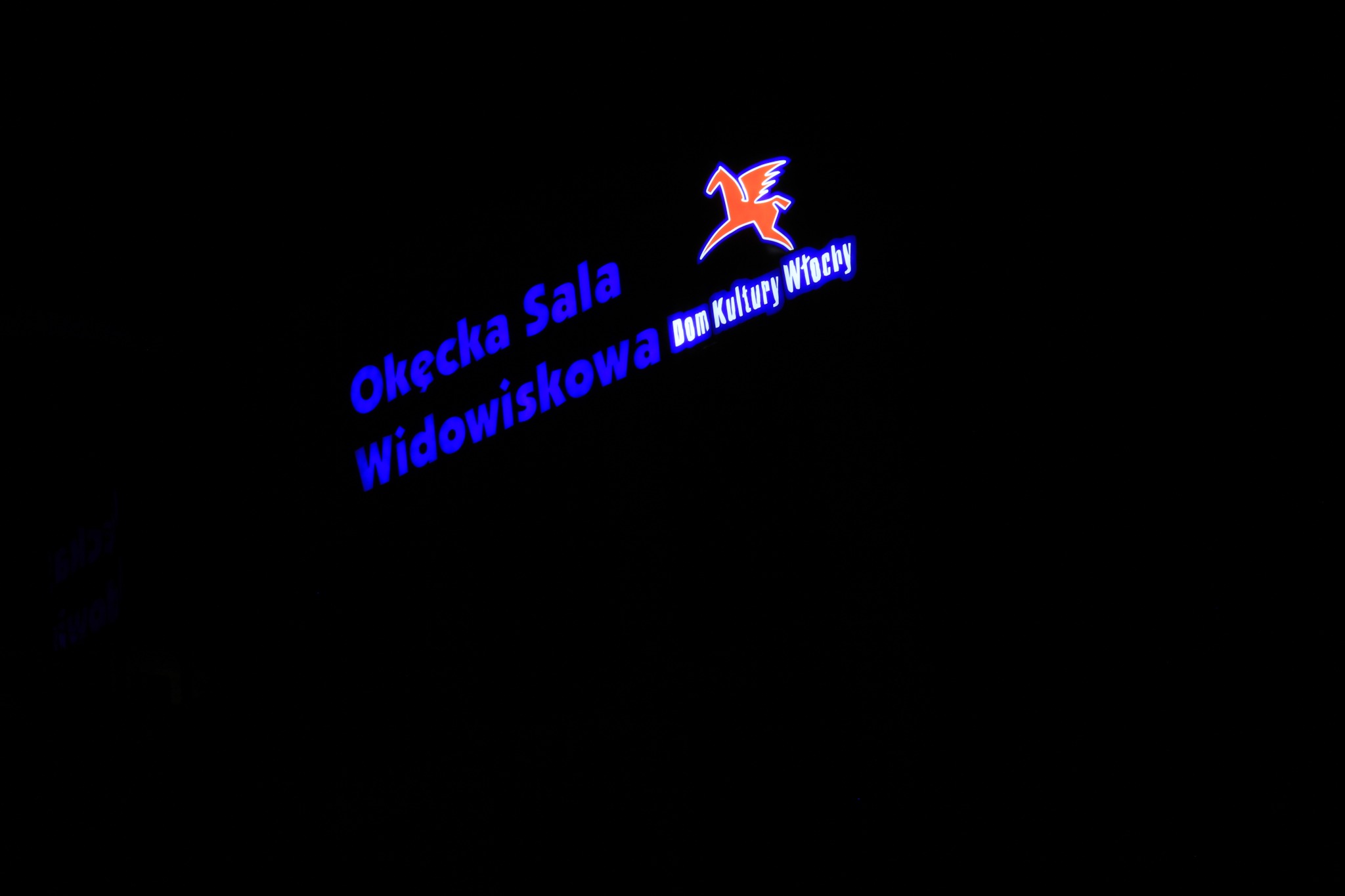 Neonowy szyld z nazwą 'Okęcka Sala Widowiskowa Dom Kultury Włochy' oraz symbolem konika z skrzydłami, wszystko w kolorach niebieskim i pomarańczowym na czarnym tle.