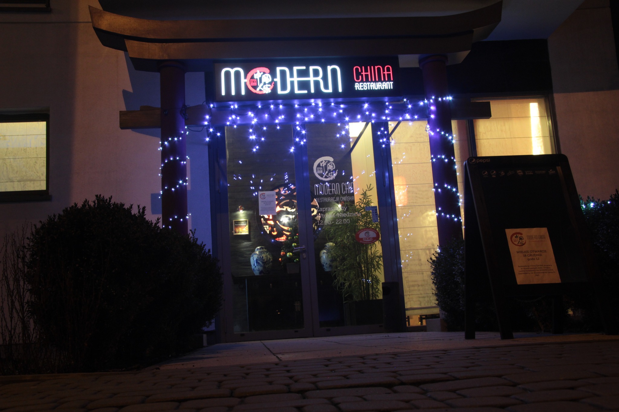 Fasada chińskiej restauracji Modern China z neonowym szyldem i dekoracyjnymi światełkami, widok z poziomu chodnika z kostki brukowej