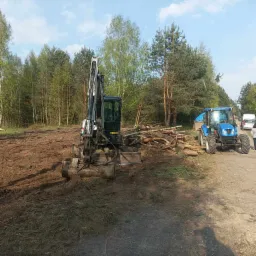 Teren po wycince drzew z widocznym mini-koparką Bobcat, stosem pociętych pni i niebieskim traktorem z przyczepą w tle.