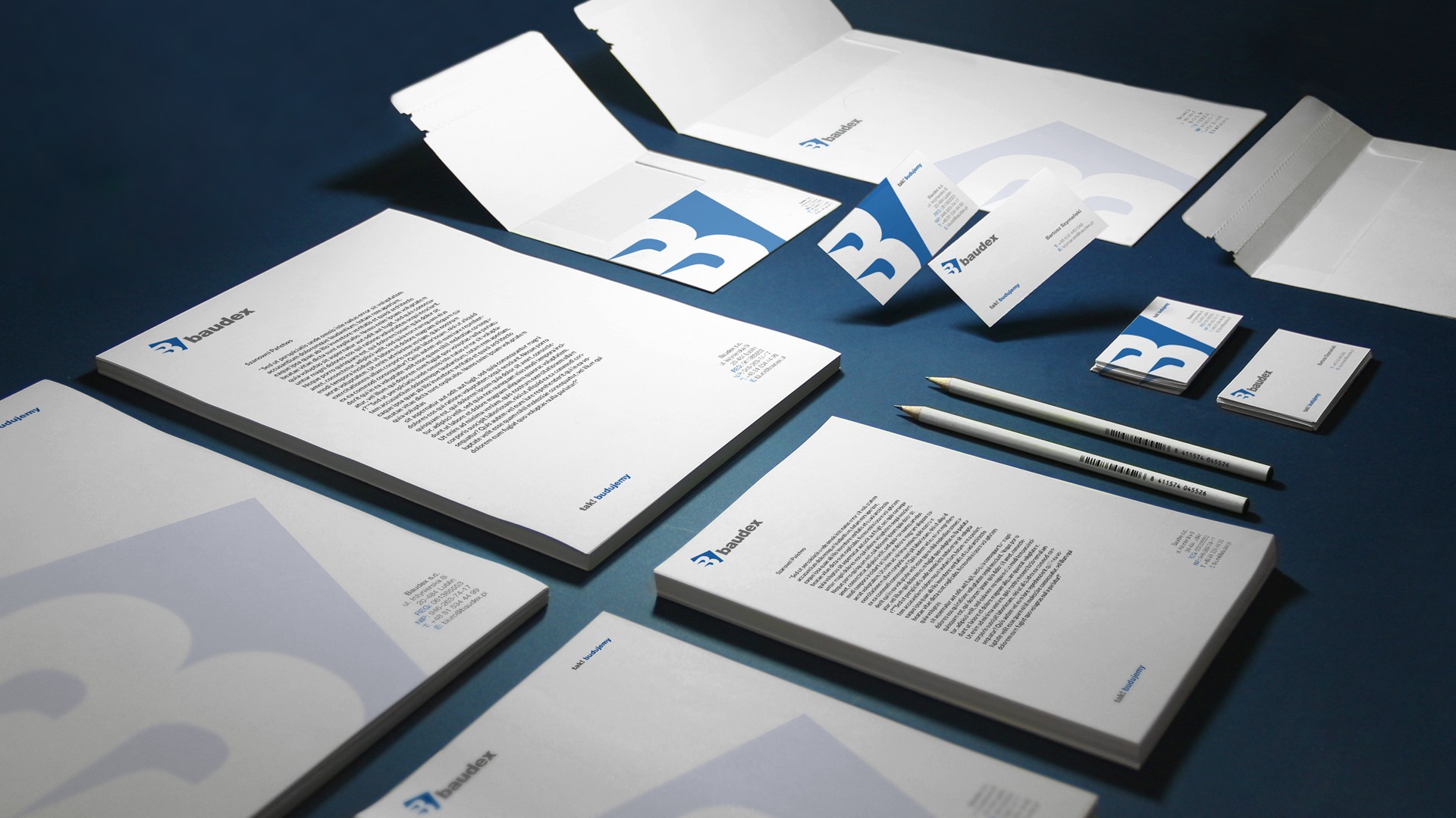 Identyfikacja wizualna firmy Baudex: papier firmowy z tekstem, koperty i wizytówki z logo na ciemnoniebieskim tle, uzupełnione dwoma ołówkami.