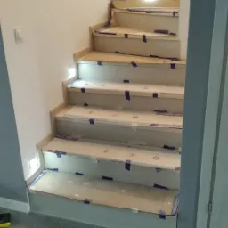 Schody wewnętrzne w trakcie montażu, zabezpieczone kartonem i taśmą malarską, widoczne oświetlenie punktowe w ścianie.