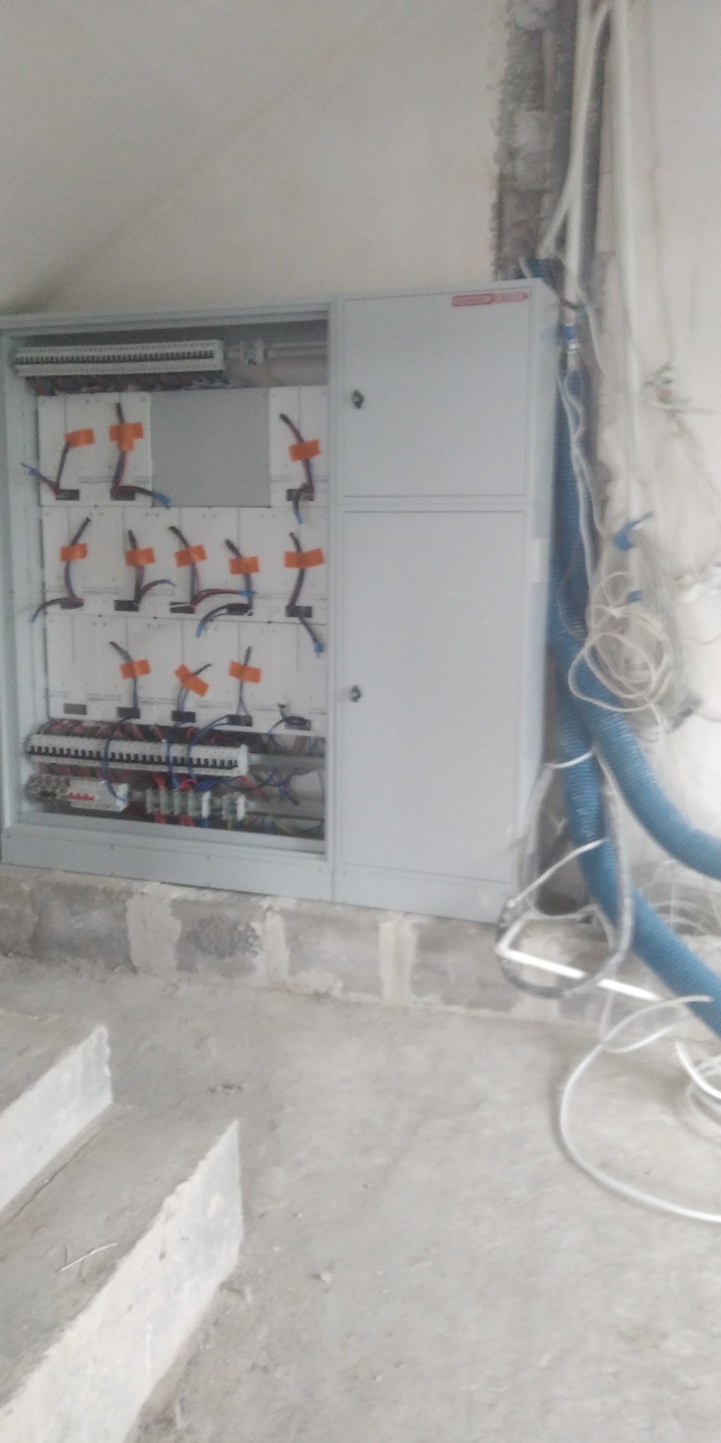 Otwarta skrzynka elektryczna z zabezpieczeniami i przewodami w surowym pomieszczeniu, niebieski karbowany peszel i luźne przewody zwisają ze ściany.