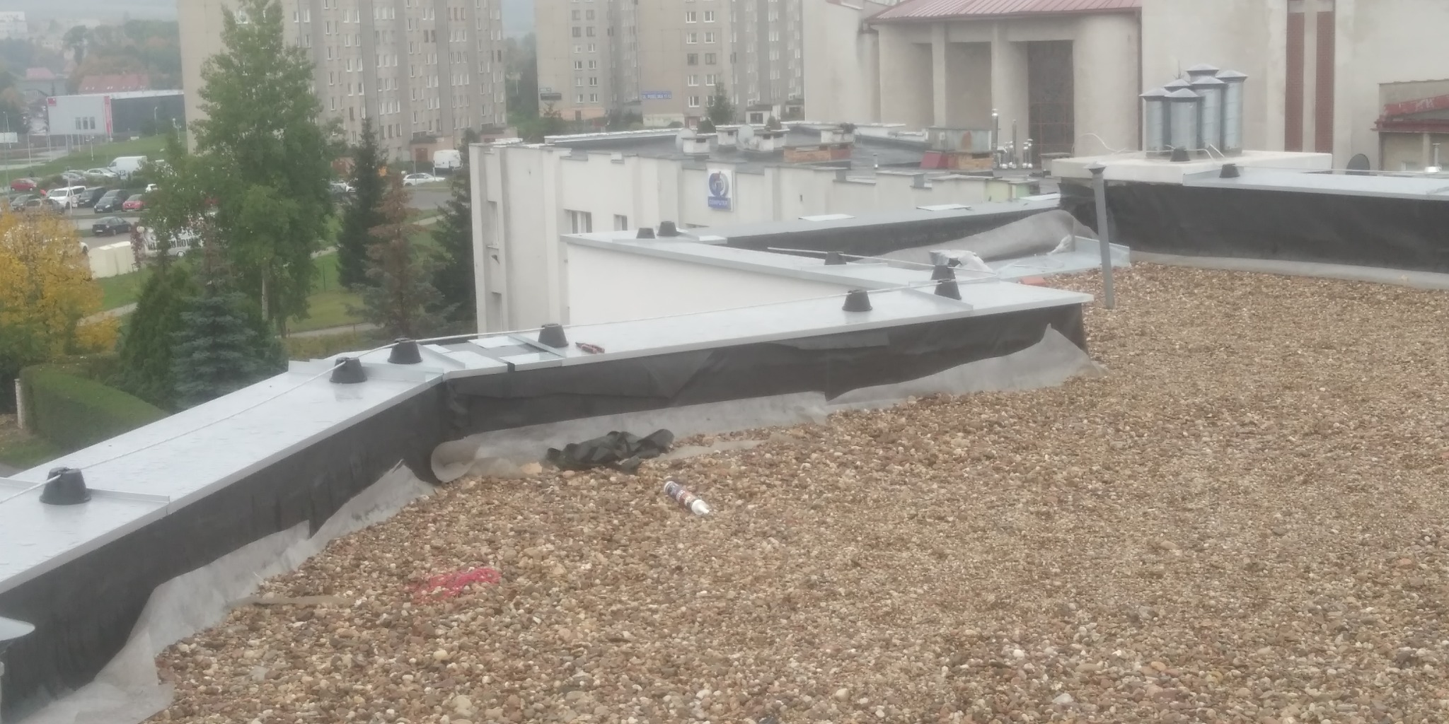 Widok z góry na płaski dach pokryty drobnym żwirem, z zamontowanymi elementami instalacji odgromowej i widocznymi budynkami w tle w pochmurny dzień.