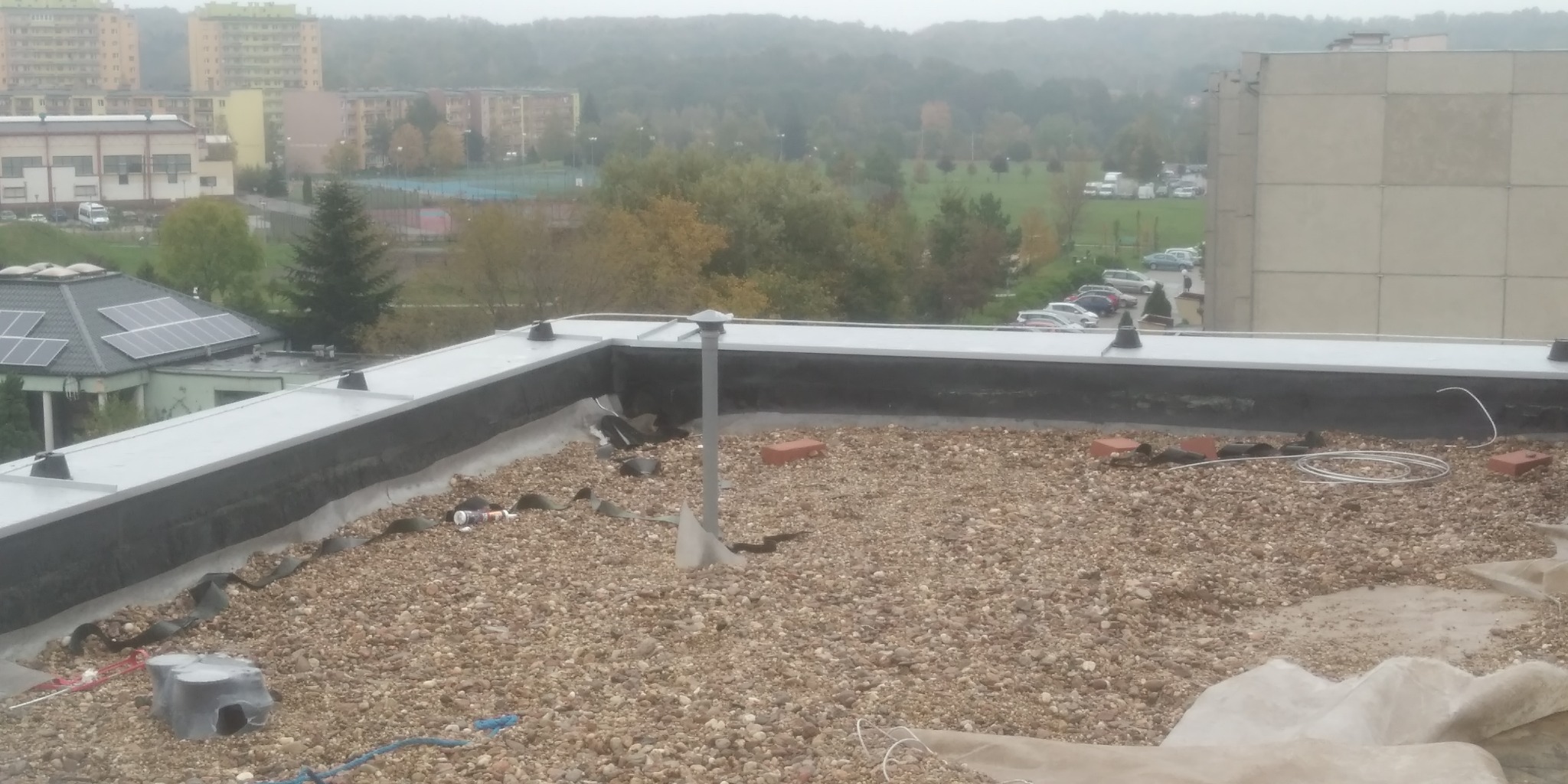 Widok z góry na płaski dach pokryty drobnym żwirem, z widocznymi elementami instalacji odgromowej w postaci metalowych wsporników i przewodów, oraz elementami instalacji elektrycznej, na tle...