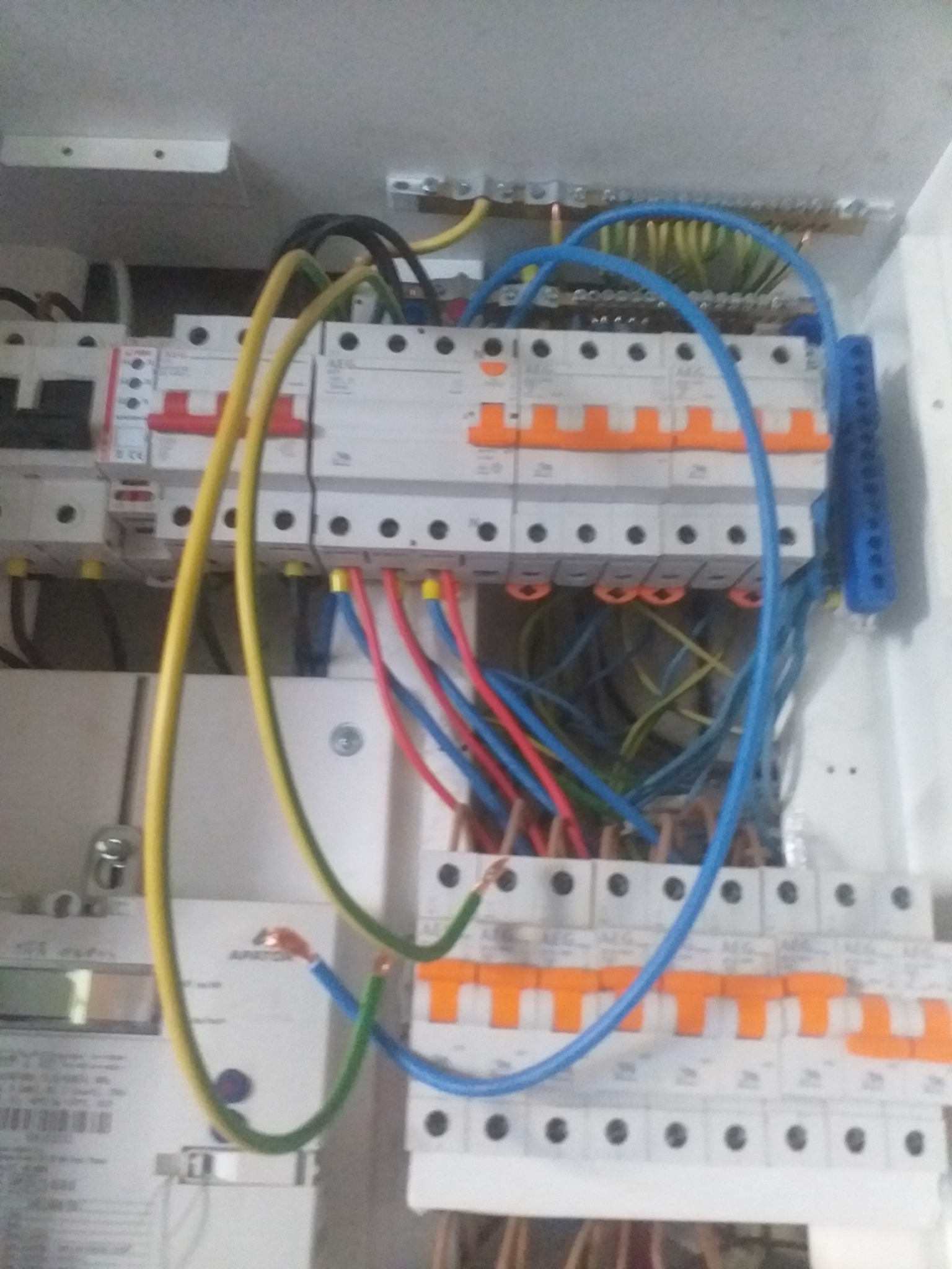 Tablica rozdzielcza z przewodami w kolorach niebieskim, czerwonym, żółto-zielonym i czarnym, podłączona do bezpieczników i licznika energii elektrycznej.