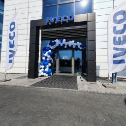 Imprezowe Niebo - Wejście do budynku z logo firmy Iveco, udekorowane łukiem z białych i niebieskich balonów, z flagami firmowymi po bokach.