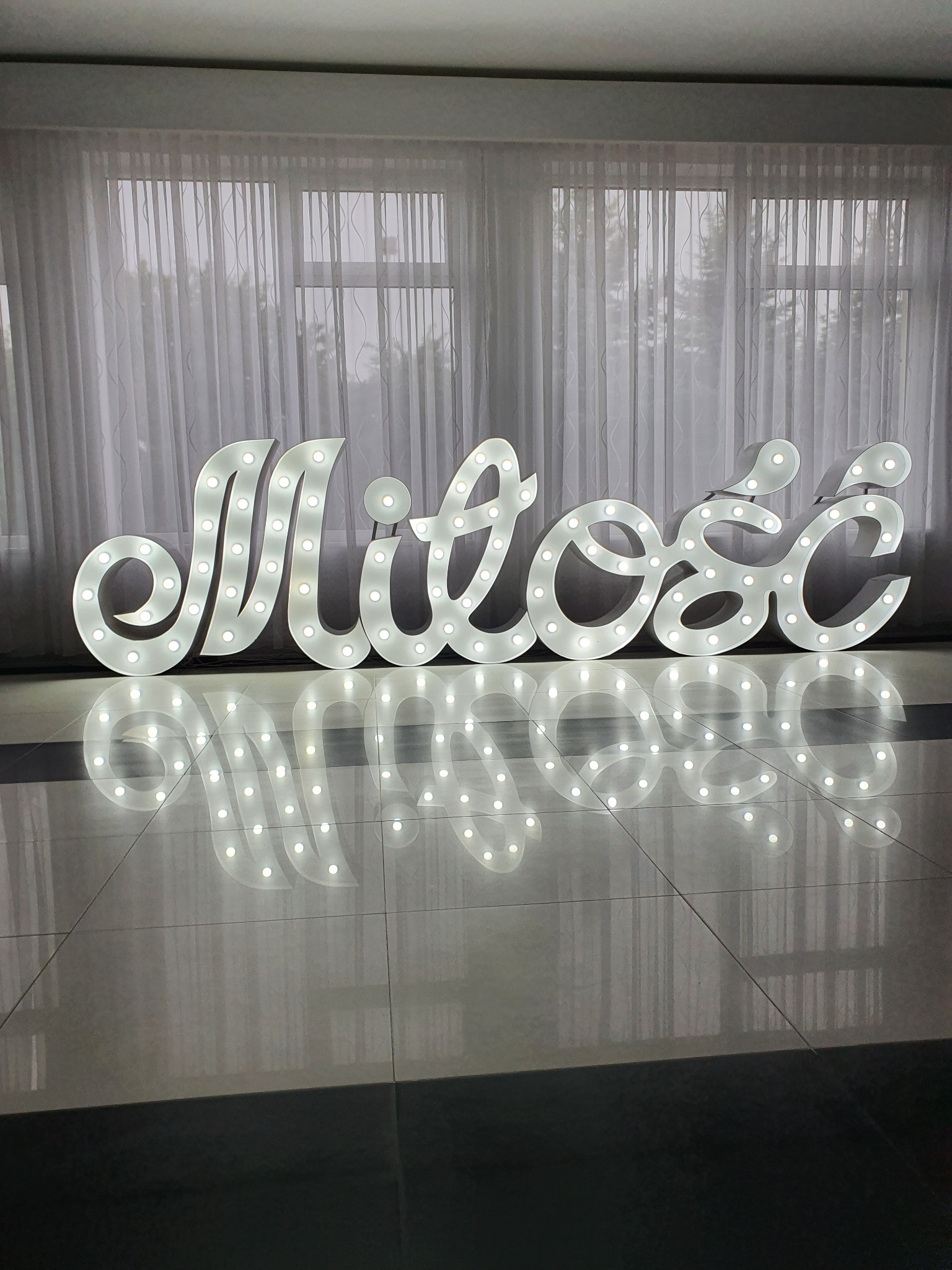 Świecący napis 'Miłość' z żarówkami na błyszczącej podłodze, odbijający się w niej, na tle okna z białymi zasłonami.