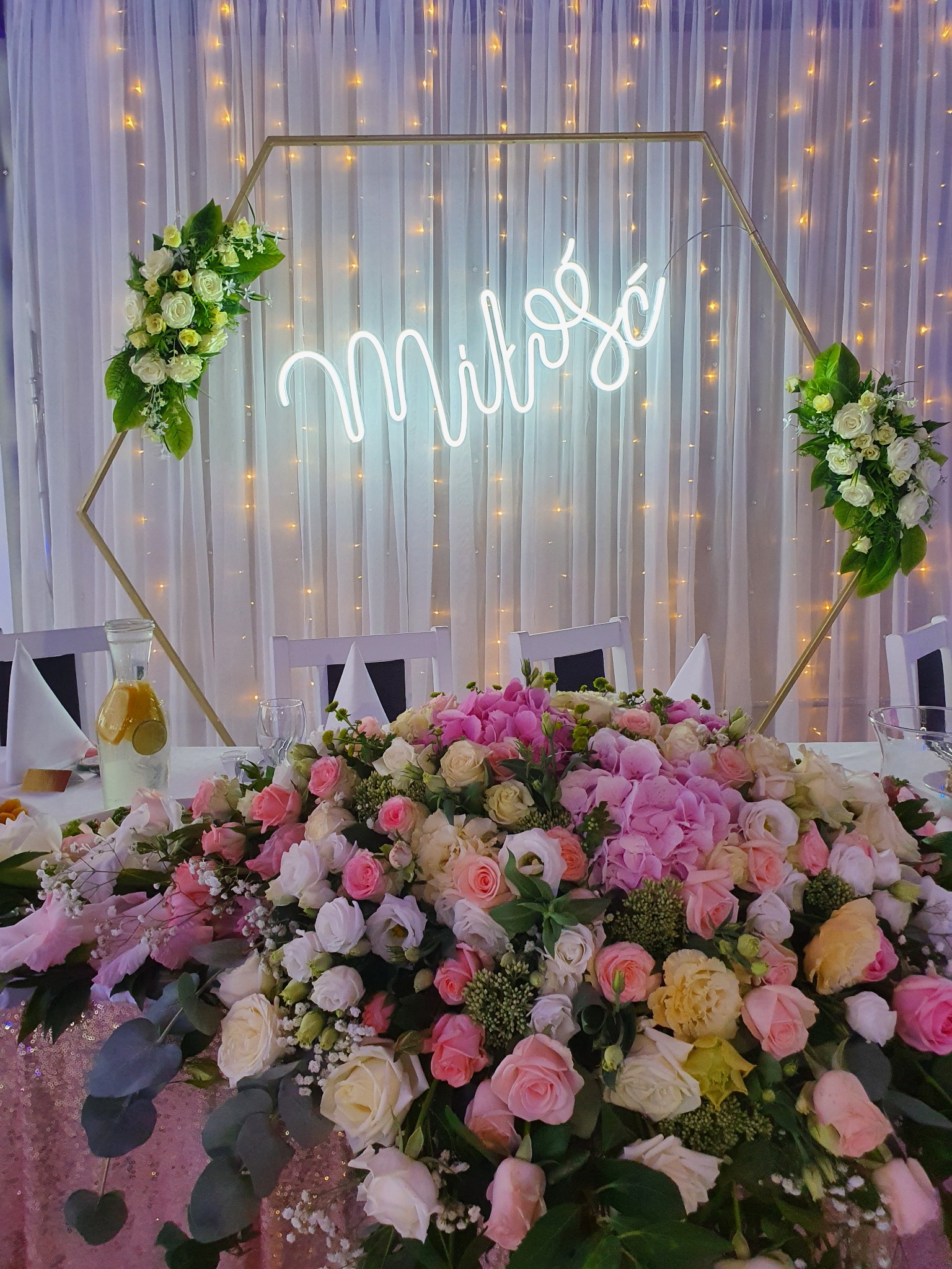 Dekoracja stołu weselnego z kompozycją kwiatową w odcieniach różu i bieli, neonowym napisem 'Miłość' w geometrycznej ramie i subtelnym oświetleniem kurtynowym w tle.