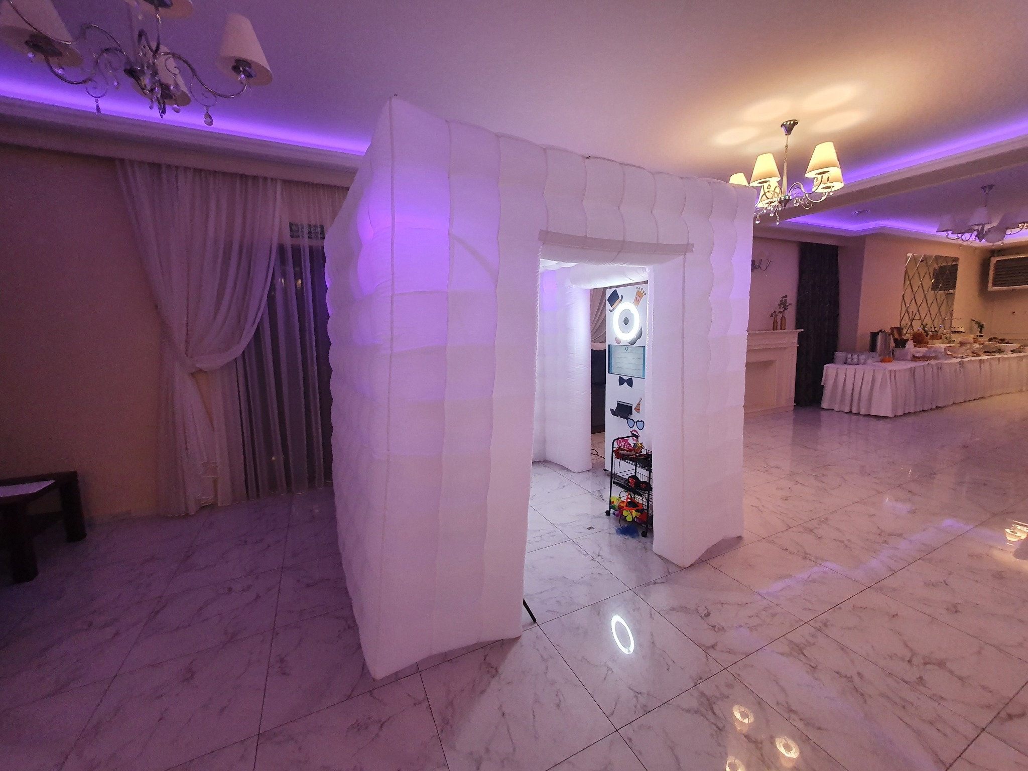 Rozstawiona biała, dmuchana fotobudka z rekwizytami wewnątrz, widoczna w sali balowej z marmurową podłogą i oświetleniem w odcieniach fioletu.