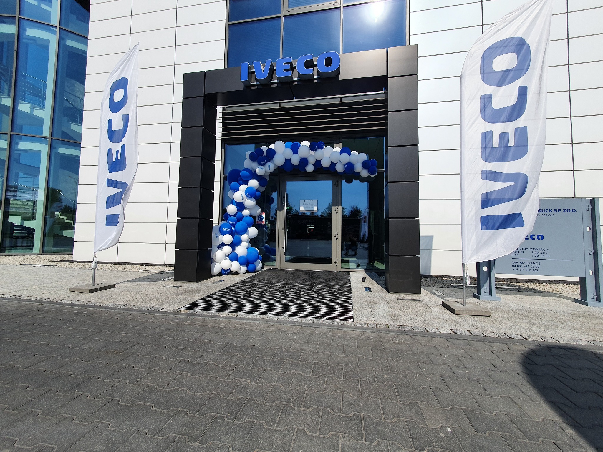 Wejście do budynku z logo firmy Iveco, udekorowane łukiem z białych i niebieskich balonów, z flagami firmowymi po bokach.