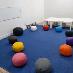 Sala lekcyjna z białymi ścianami, tablicą, niebieskim dywanem, białym stolikiem i okrągłymi, kolorowymi pufami do siedzenia, oraz dekoracjami na ścianie.
