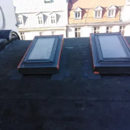 Dwa okna dachowe zamontowane na płaskim dachu pokrytym papą, widoczne budynki w tle.