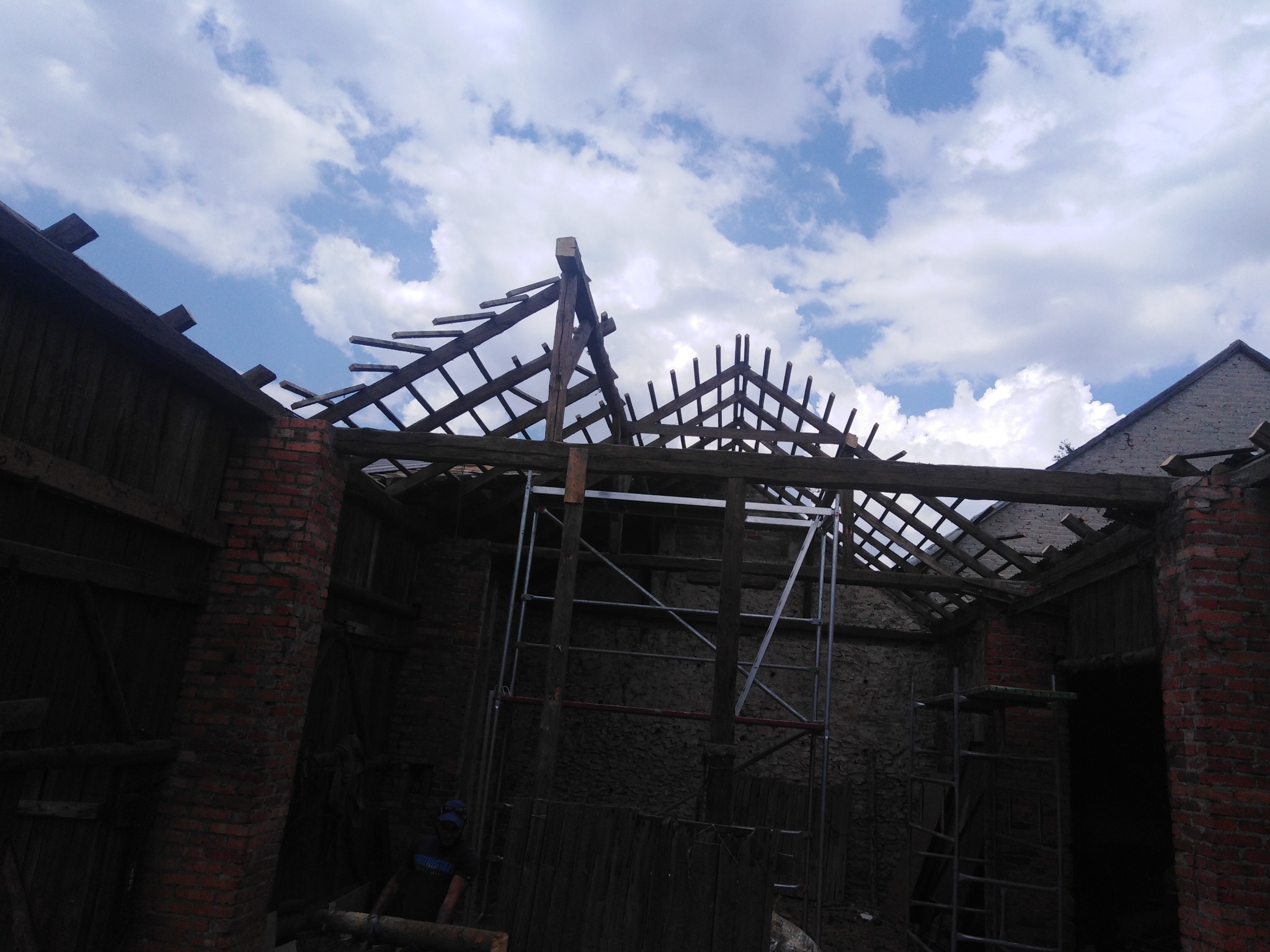 Konstrukcja drewniana dachu w trakcie budowy, widoczne krokwie i rusztowanie wewnątrz budynku z ceglanymi ścianami, na tle błękitnego nieba z chmurami.