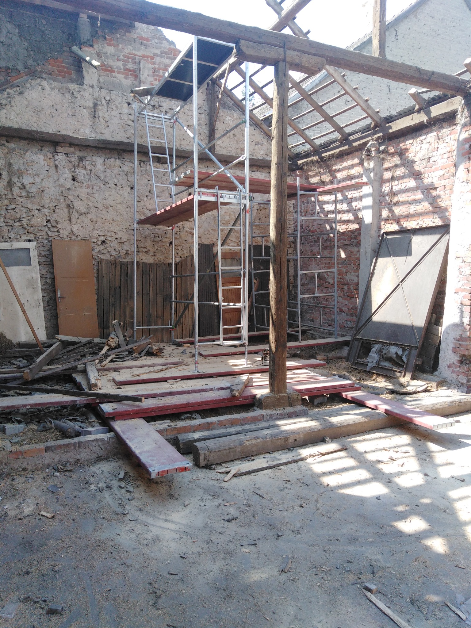 Wnętrze budynku w trakcie remontu dachu, widoczne rusztowanie, fragmenty starej konstrukcji drewnianej i ceglanej, gruz i elementy demontażu na podłożu.