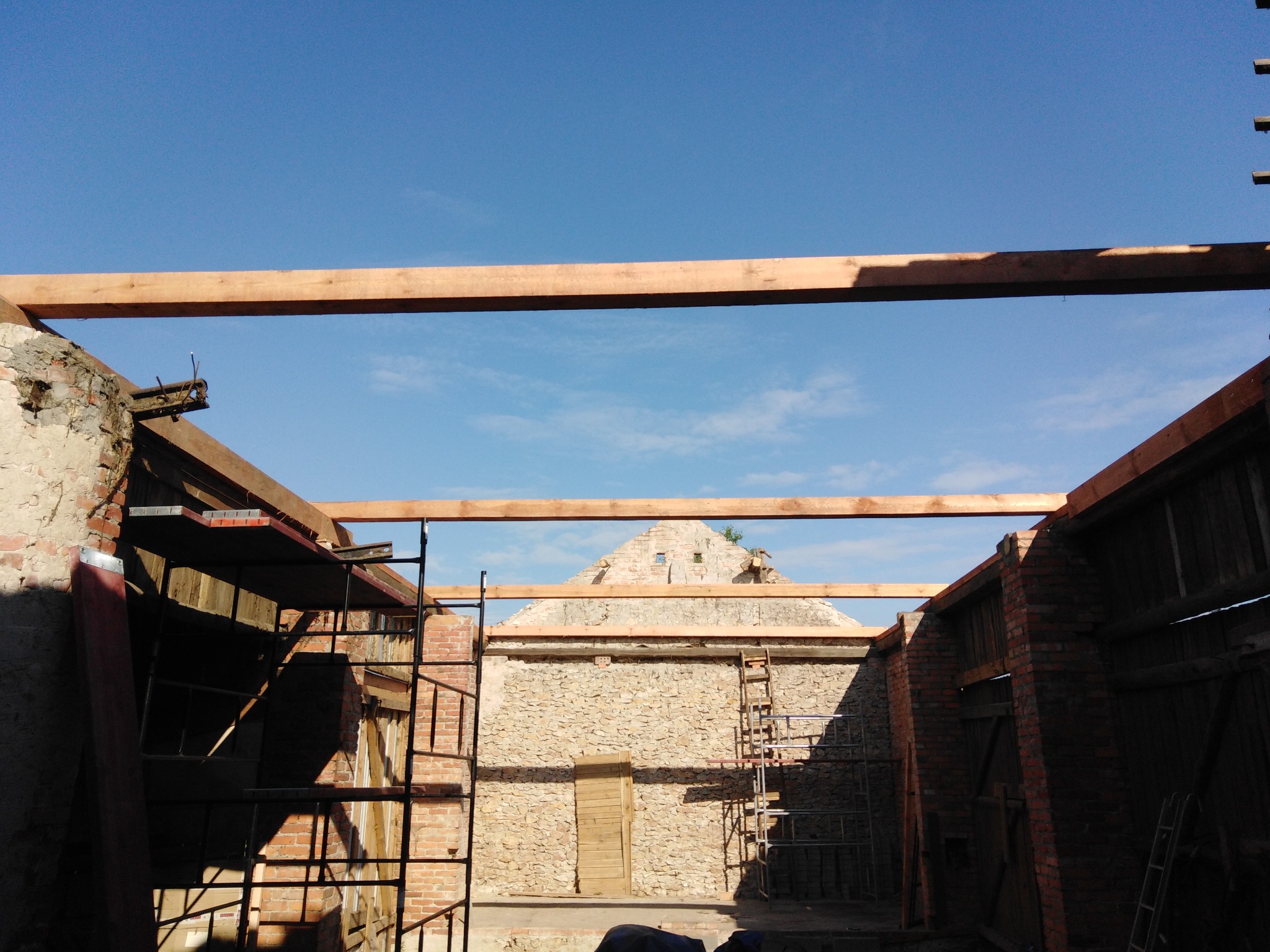 Montaż drewnianej konstrukcji dachu wewnątrz budynku z kamiennymi ścianami, widoczne rusztowania i fragmenty starego muru.