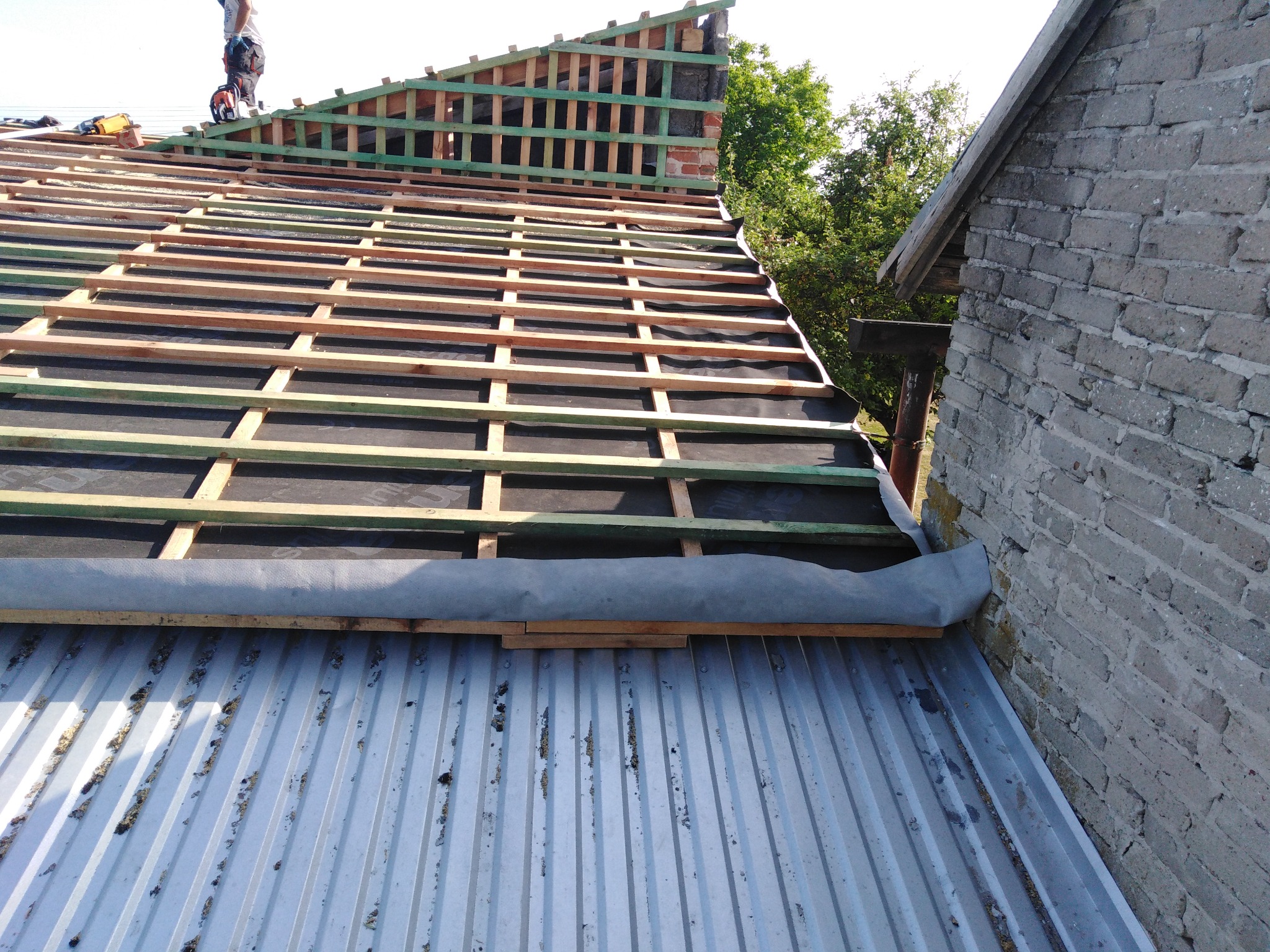 Konstrukcja dachu z drewnianymi łatami i membraną hydroizolacyjną, widok z góry na blaszane pokrycie z zabrudzeniami, dekarz na szczycie dachu z pilarką.