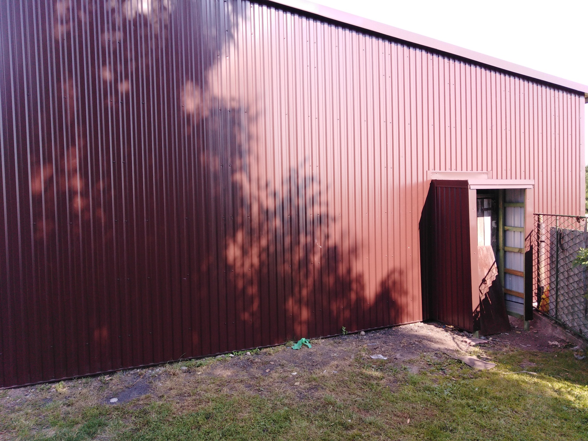 Brązowa blaszana ściana budynku gospodarczego z widocznym wejściem i cieniem drzewa.