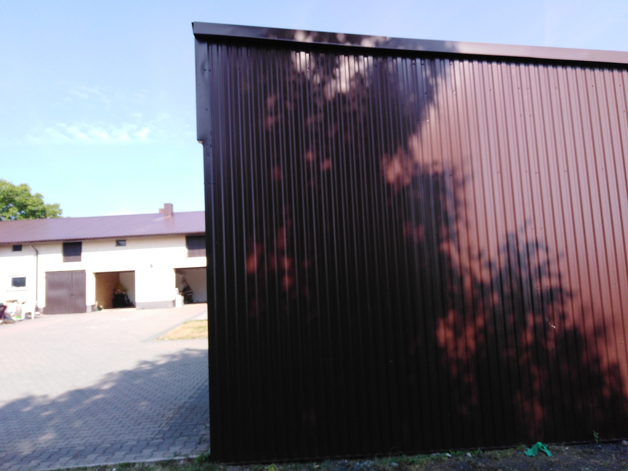 Brązowa, blaszana ściana budynku gospodarczego z widocznymi cieniami drzew, w tle budynek mieszkalny z brązowym dachem.