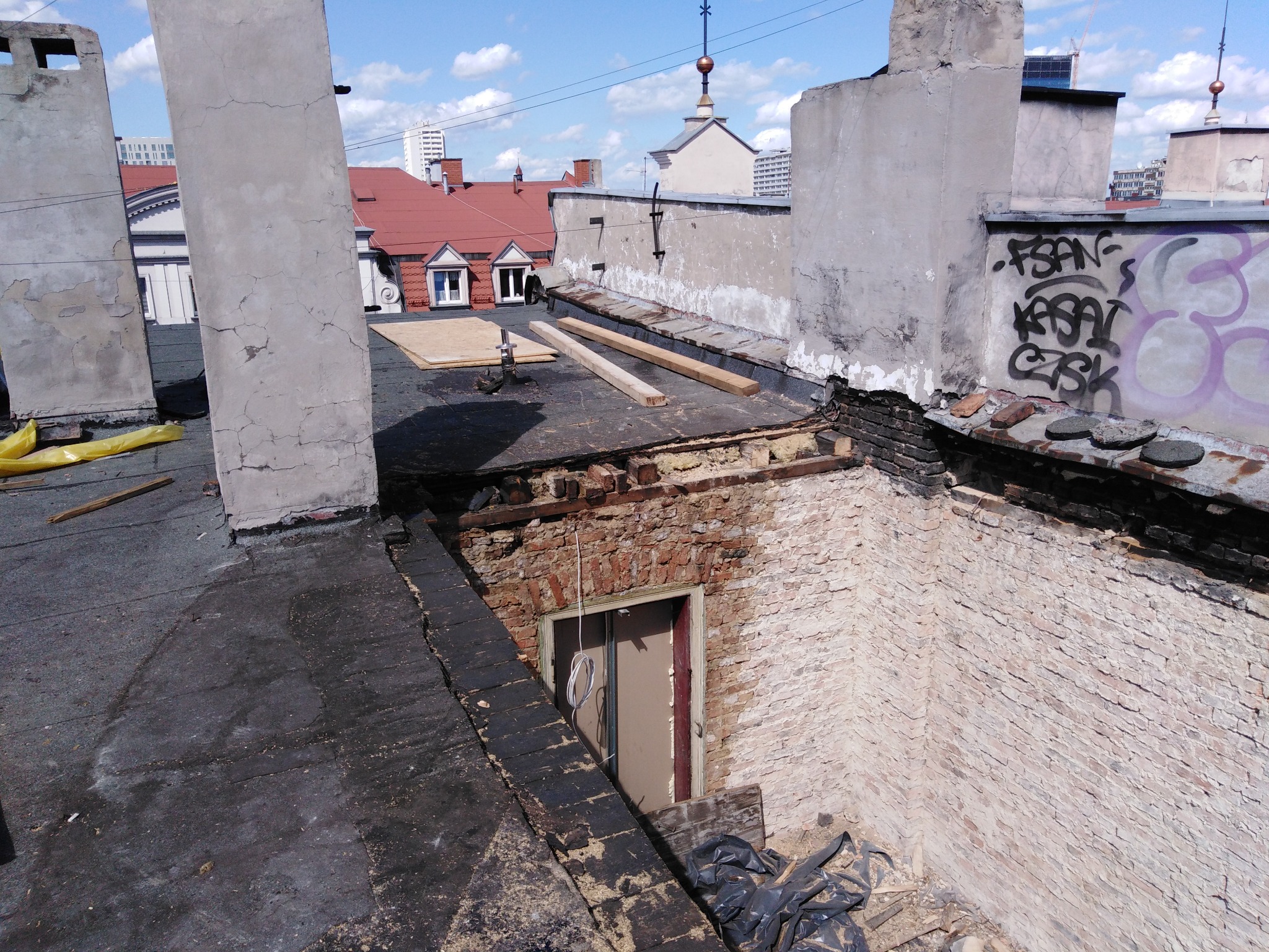 Uszkodzony dach budynku w trakcie renowacji, widoczne ceglane kominy, odsłonięta konstrukcja i materiały budowlane, w tle panorama miasta.