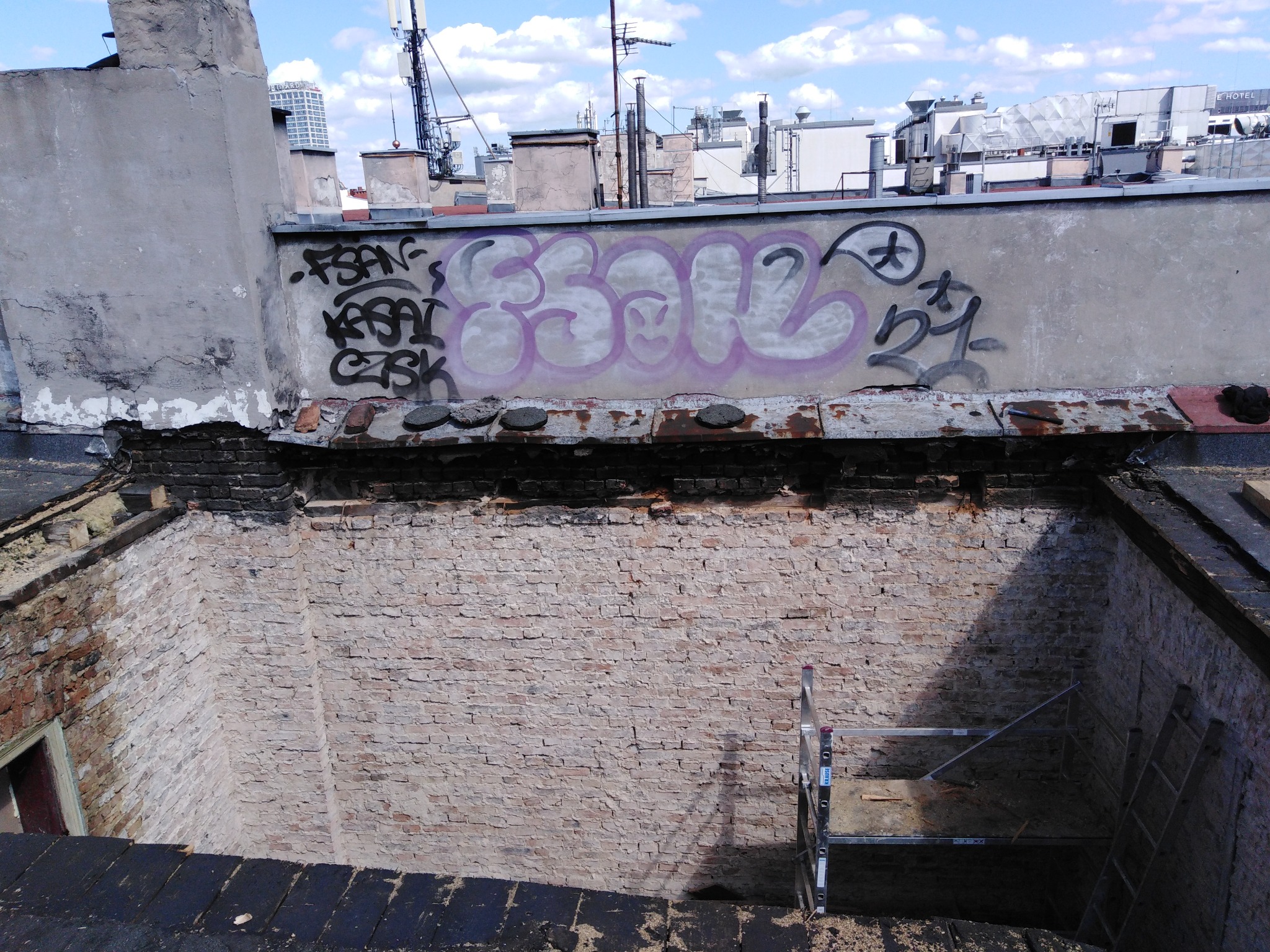 Odsłonięta konstrukcja dachu z ceglaną wnęką, widoczne graffiti na ścianie, drabina aluminiowa wewnątrz, panorama miejska w tle.