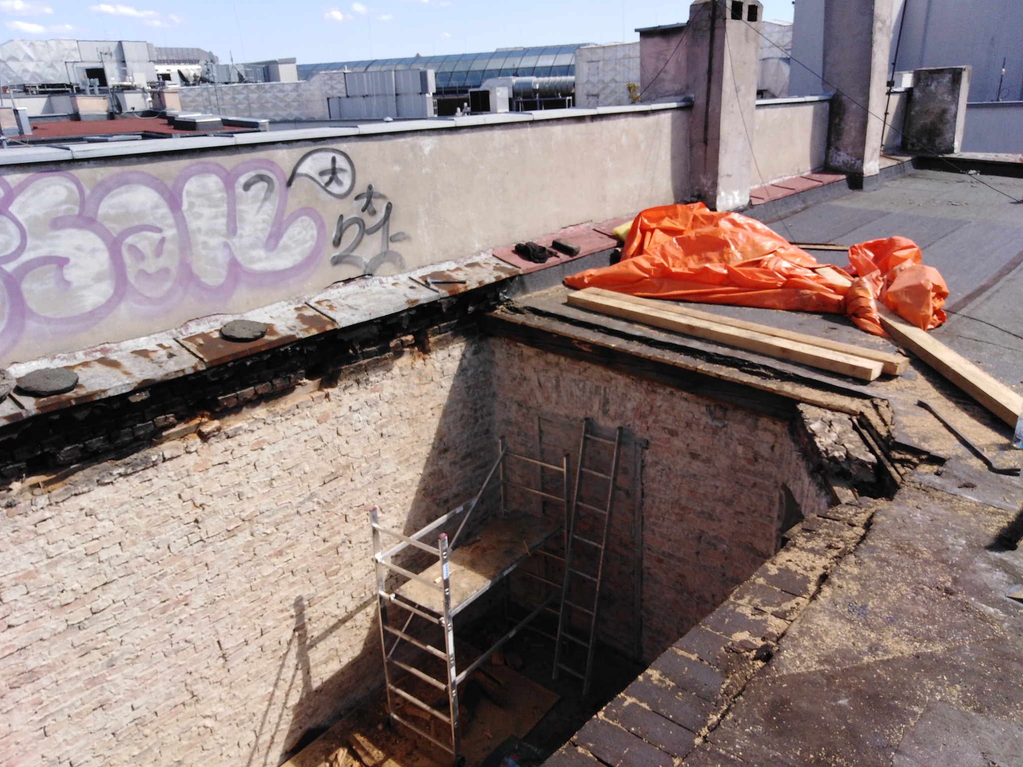 Odkryty fragment dachu z ceglaną ścianą komina, widoczne rusztowanie wewnątrz, zabezpieczone pomarańczową plandeką, graffiti na ścianie budynku w tle.