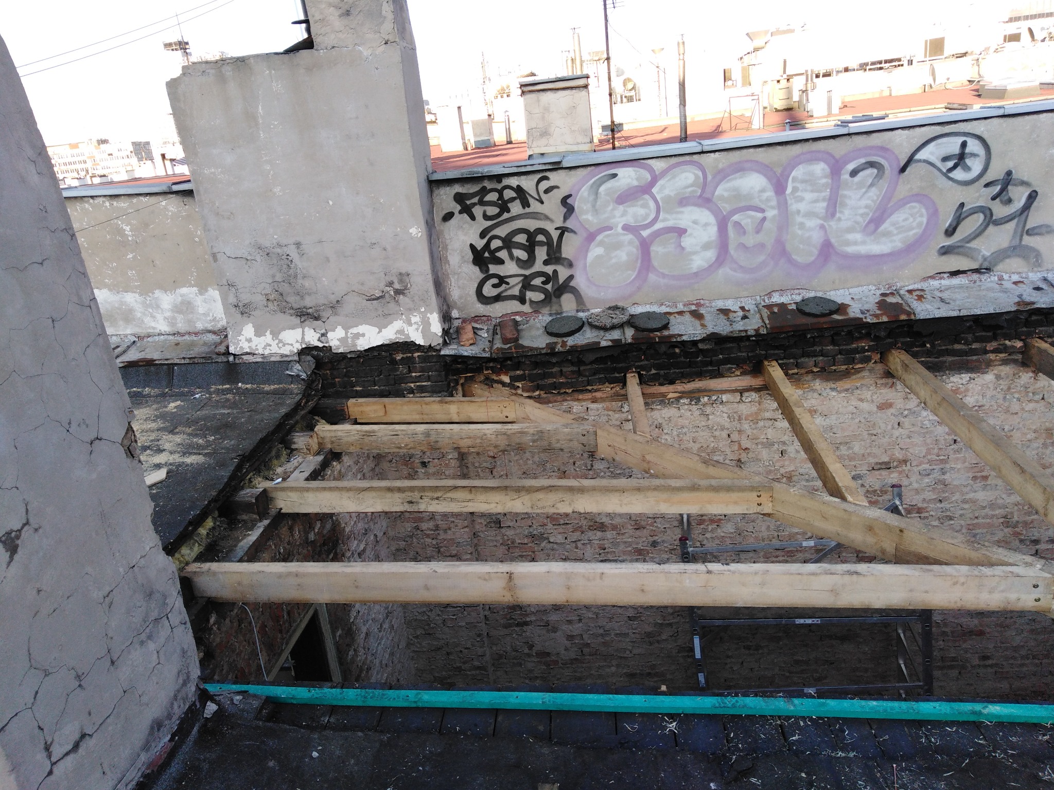 Konstrukcja więźby dachowej z drewnianych belek na tle ceglanej ściany i graffiti na dachu kamienicy.