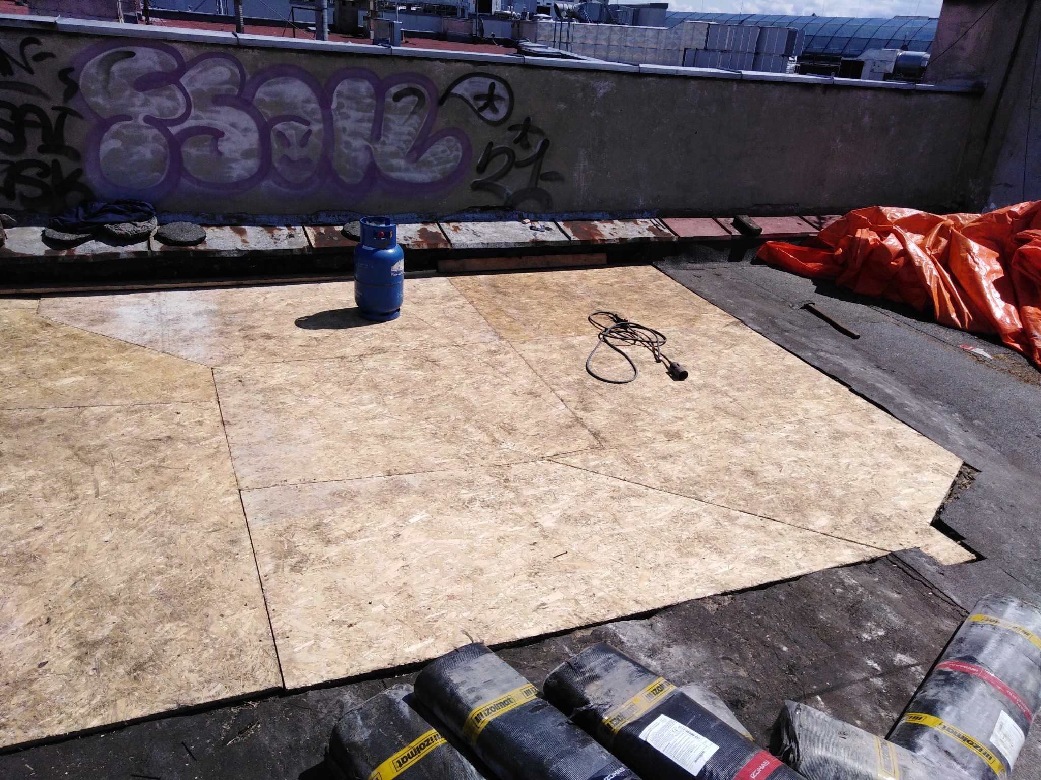 Przygotowanie dachu: płyty OSB ułożone na bitumicznej powierzchni, butla gazowa, zwinięty kabel grzewczy oraz rolki papy bitumicznej w dolnej części kadru, ściana z graffiti w tle.
