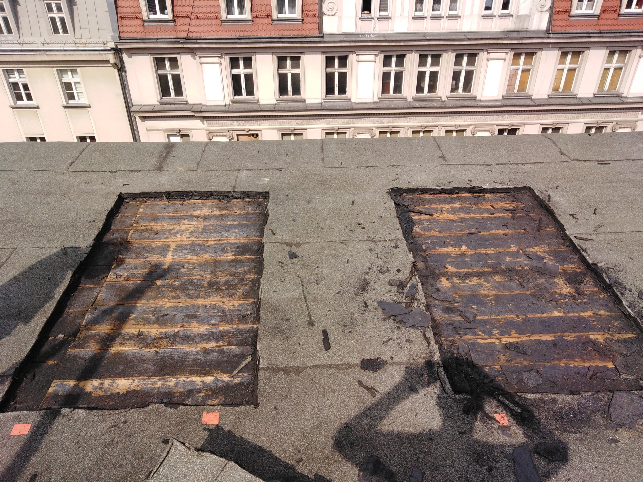 Fragment dachu z widocznymi dwoma uszkodzonymi fragmentami poszycia, odsłaniającymi drewnianą konstrukcję, na tle budynków mieszkalnych.