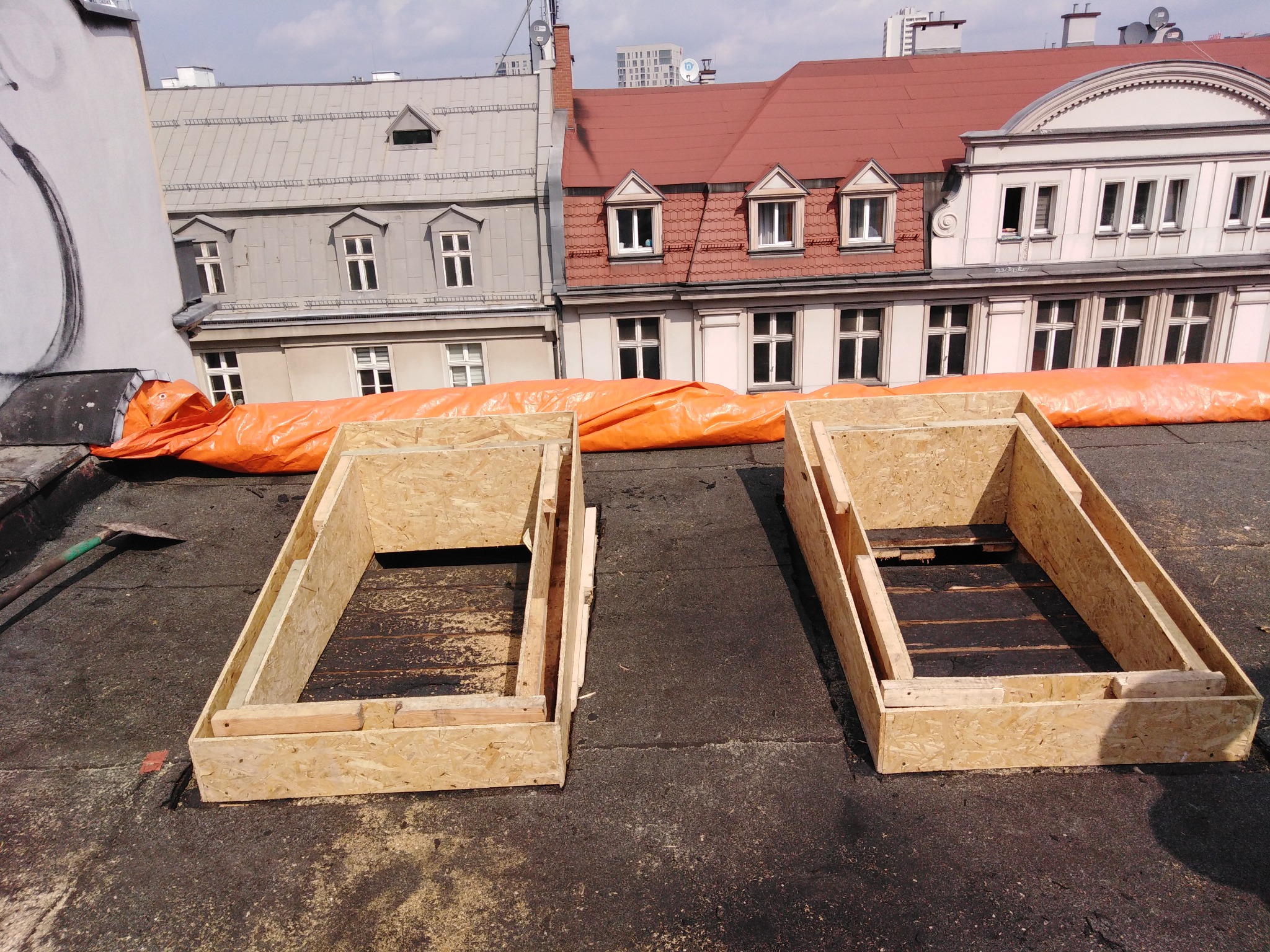 Dwa drewniane szalunki na płaskim dachu pokrytym papą, w tle budynki mieszkalne z różnymi pokryciami dachowymi i pomarańczowa plandeka zabezpieczająca.