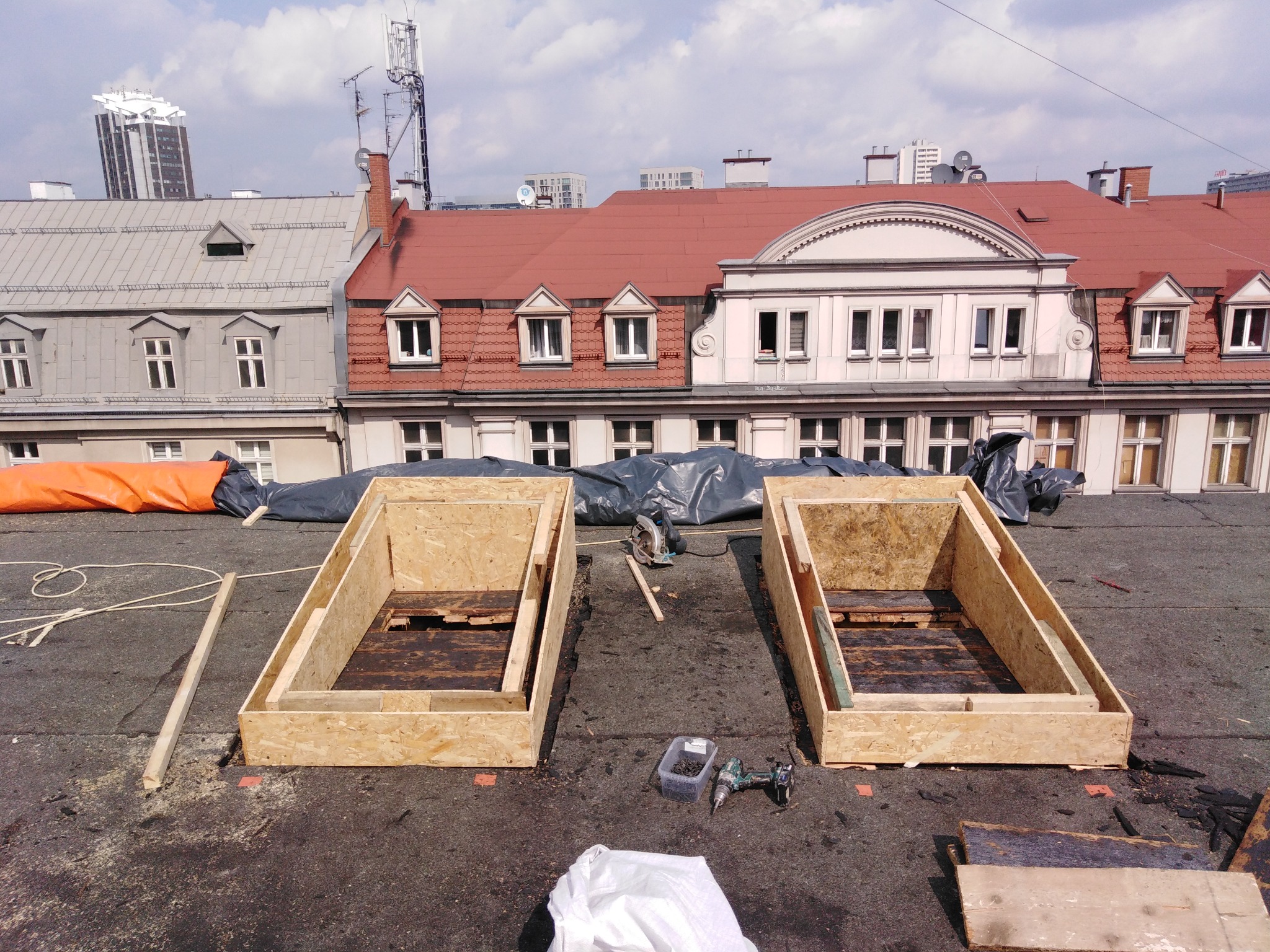 Dwa drewniane szalunki na płaskim dachu budynku w trakcie budowy lub remontu, narzędzia i materiały budowlane w pobliżu, zabudowa miejska w tle.